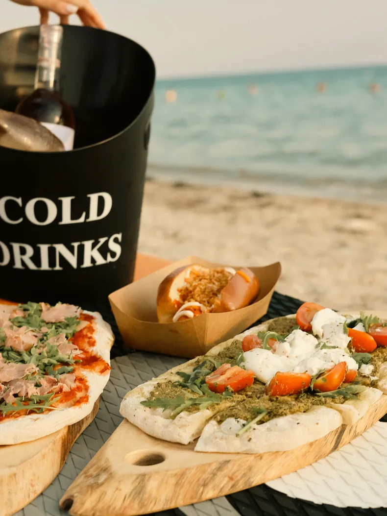 Flatbreads and a hot dog served on the beach next to a cold drinks bucket